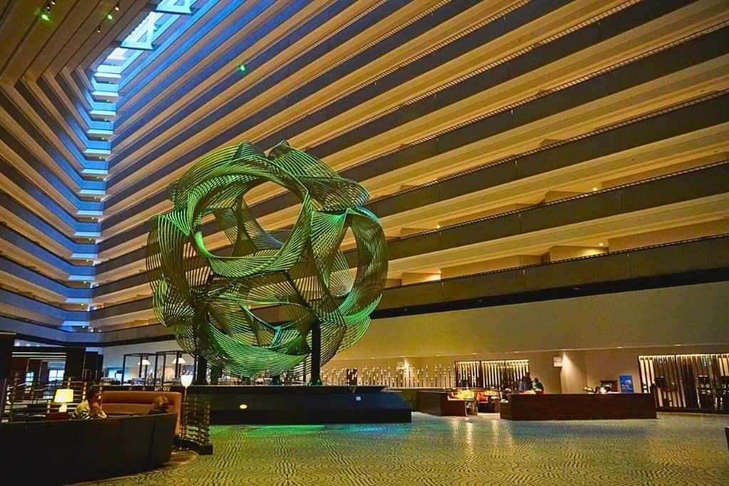 An interior view of a hotel lobby with a large, intricate green sculpture as the centerpiece. The lobby is spacious with contemporary furnishings and a tiered, multi-level ceiling that adds depth to the space.