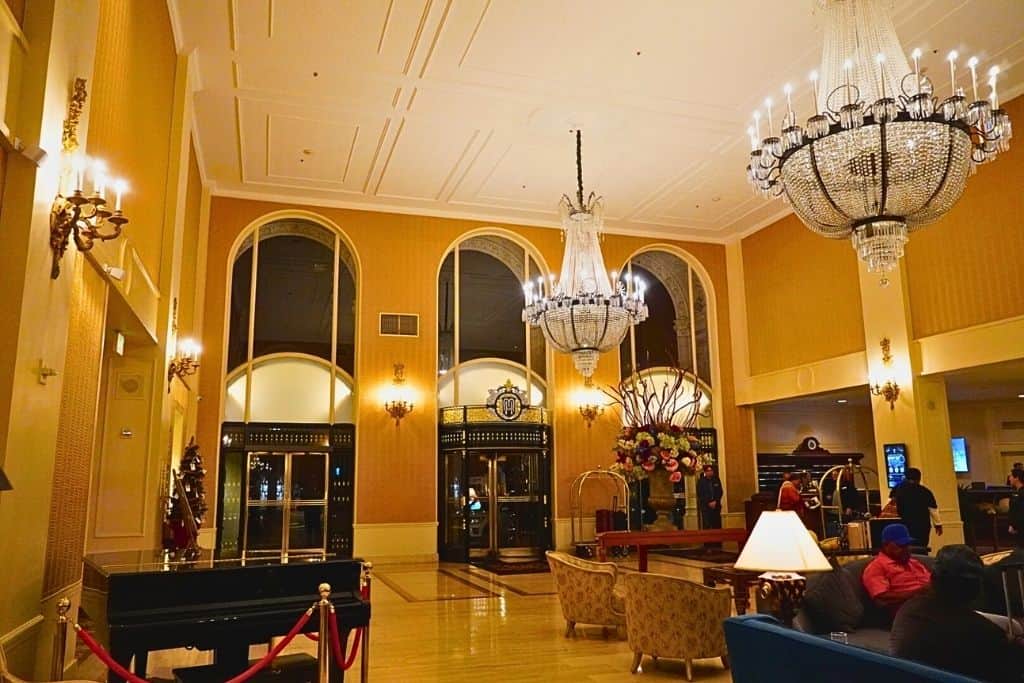 A grand hotel lobby rich in traditional elegance, featuring 2 large, ornate chandeliers, luxurious floral centerpiece, and classic furnishings, with guests and staff adding a lively ambiance.