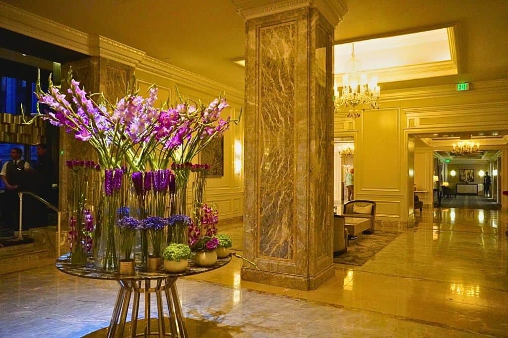 An elegant hotel lobby interior, adorned with tall vases of vibrant purple flowers and greenery on a round table, with luxurious marble columns and crystal chandeliers enhancing the opulent setting.