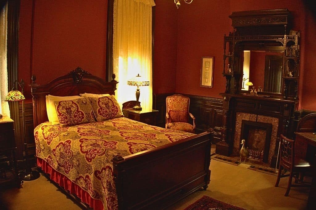 An intimate bedroom setting featuring a luxurious bed with patterned bedding, antique wood furniture, and decorative lamps, providing a sense of Victorian elegance and comfort.