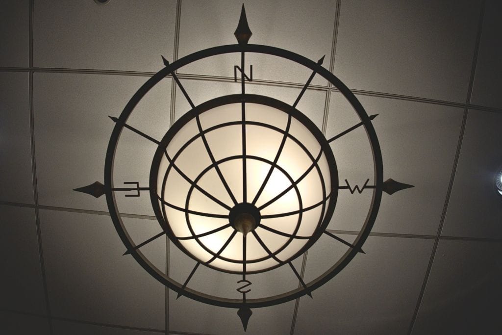 A compass ceiling light fixture, with the cardinal directions marked by arrows pointing north, south, east, and west, illuminating softly against a drop ceiling.