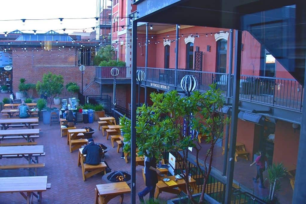 An outdoor seating area of a restaurant or bar with wooden tables and benches, string lights above, and a view of balconies with the sign "San Francisco Brewing Co."