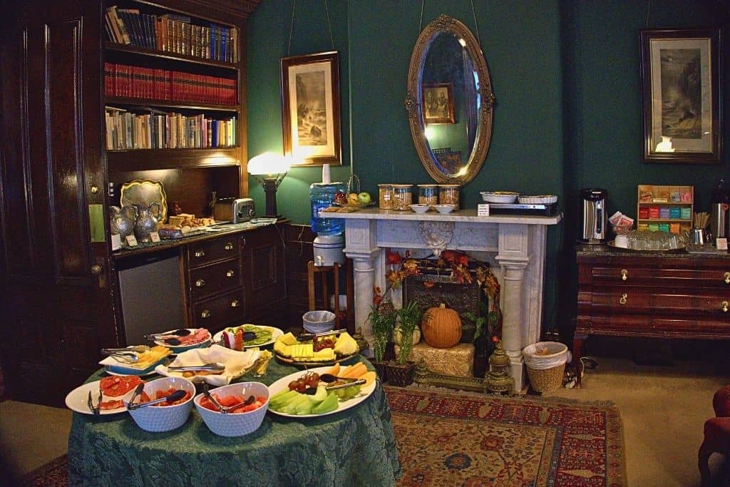 A cozy and traditional dining space with a table set with an array of breakfast foods, flanked by a marble fireplace and a bookshelf filled with volumes, conveying a homey and inviting atmosphere.