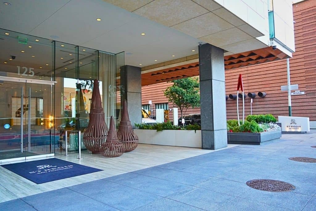The entrance of a luxurious hotel with large glass doors flanked by striking column-like sculptures and a welcoming mat, set against a backdrop of polished stone and a neatly landscaped planter, inviting guests into an atmosphere of exclusivity and comfort.