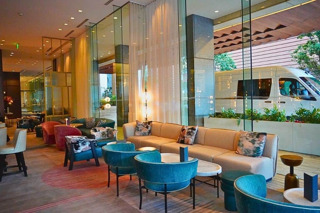 A sophisticated hotel lounge featuring an elegant mix of plush seating arrangements in pastel and deep teal hues, accented by decorative pillows, with a warm, inviting glow from stylish wall-mounted lights reflecting off the glass partitions, creating a serene and fashionable space for guests to unwind.