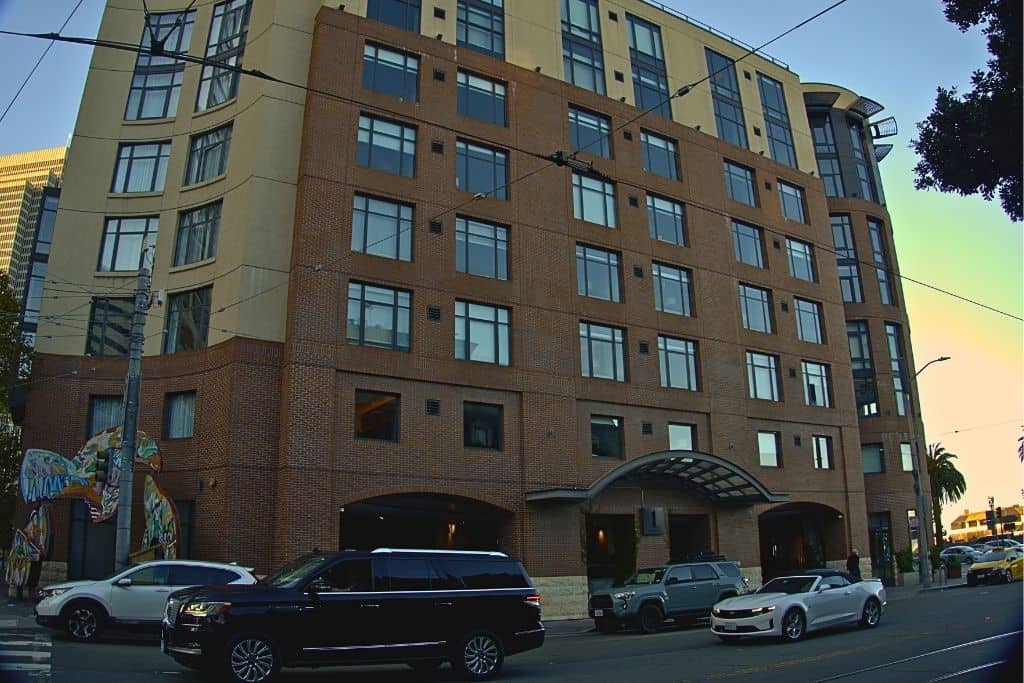 A large, multi-story building with a brick facade on a street corner. The building features rows of windows and an arched entrance with a canopy overhead. Cars and a painted mural are visible along the street.
