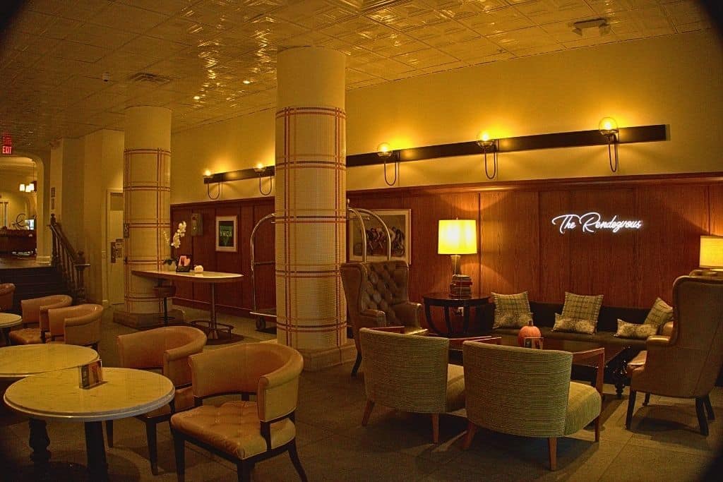 Inside a cozy lounge named "The Rendezvous", there are comfortable chairs and sofas with pillows, round tables, and a warm glow from wall lights. Decorative columns and a reflective ceiling add to the elegant atmosphere.