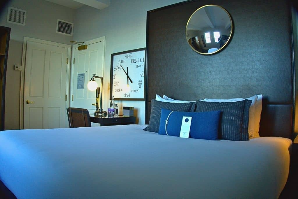 A modern hotel room with a large bed in the center, a dark gray headboard, and pillows of different sizes. A desk with a lamp and a large clock with mathematical equations on the wall make the room look both stylish and a bit like a classroom.