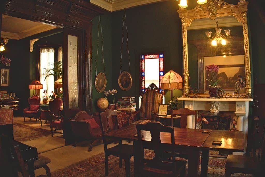 A vintage-style room with deep green walls and dark wooden trim, featuring antique furniture, a large gold-framed mirror, and stained glass windows, creating a cozy, historic atmosphere.