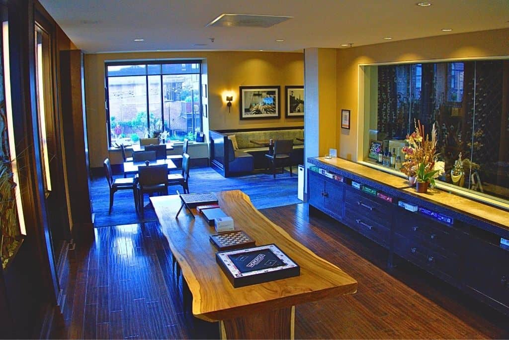 A dimly lit, sophisticated interior space of a hotel with a wooden communal table, plush seating areas, and a bar counter with games with a mirror backdrop reflecting the room.