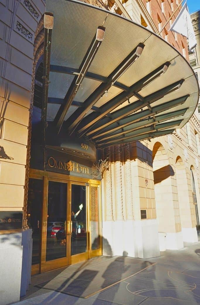 The entrance to the Omni Hotel is showcased with a classic awning extending over the sidewalk, the hotel's name in elegant gold lettering above the glass doors, and the warm glow of interior lighting against the building's stone facade.