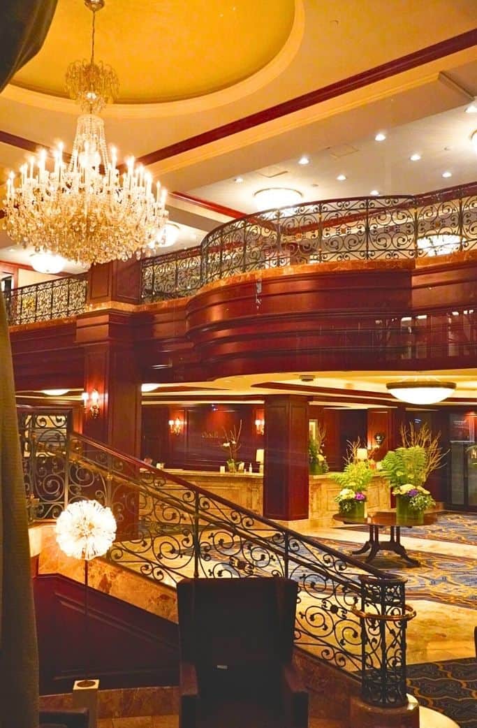 A luxurious hotel lobby featuring a grand chandelier, a sweeping staircase with ornate railings, and a warm, inviting reception area accented by plush floral arrangements and rich wood finishes.