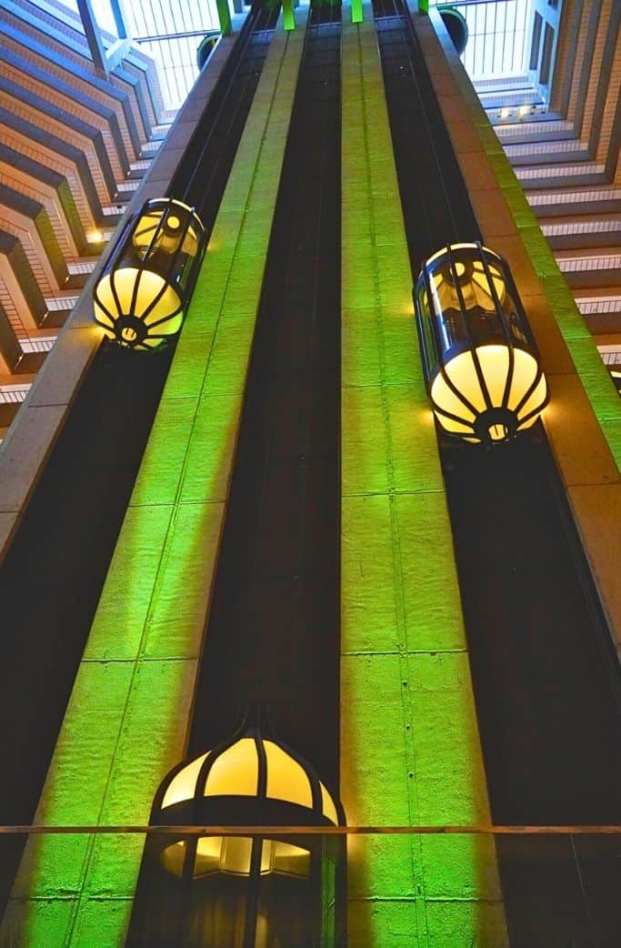 A view from the bottom of an elevator shaft inside a hotel, showing three elevators with art deco-style light fixtures illuminating the space, accentuating the vertical lines of the building's interior architecture.
