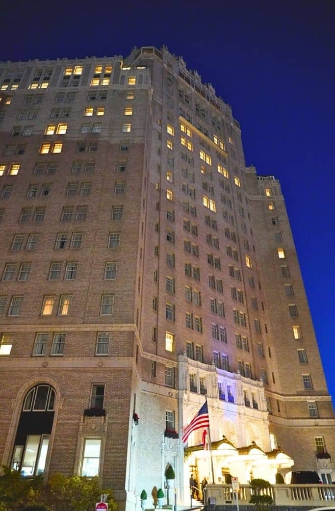 The stately exterior of a historic hotel building at night, with illuminated windows, a grand entrance, and an American flag, as seen from a street-level perspective.