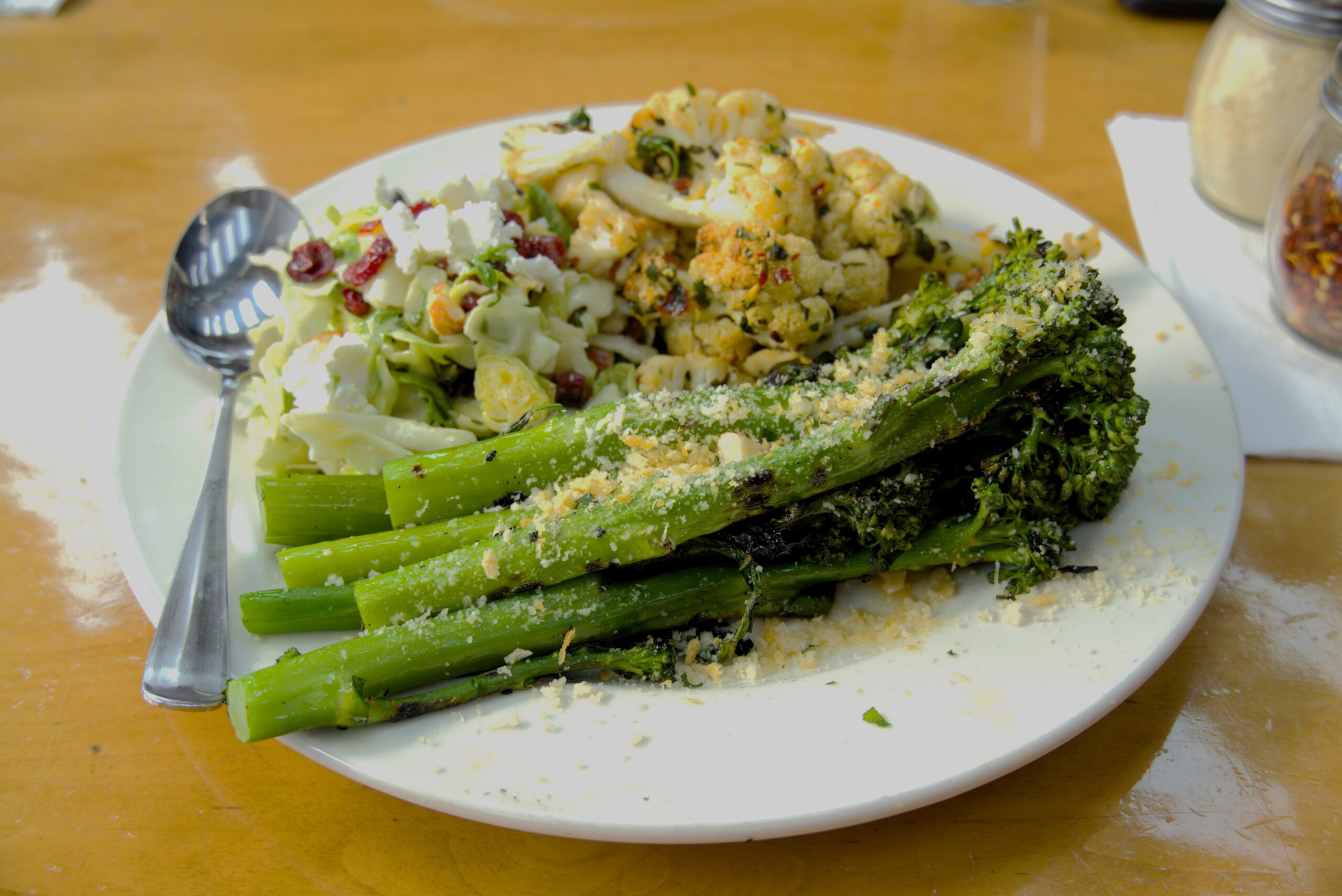 This is a picture of a plate of food on a table. It has green asparagus and broccoli, with some cauliflower salad on the side. The food looks healthy and is sprinkled with some cheese or breadcrumbs on top.
