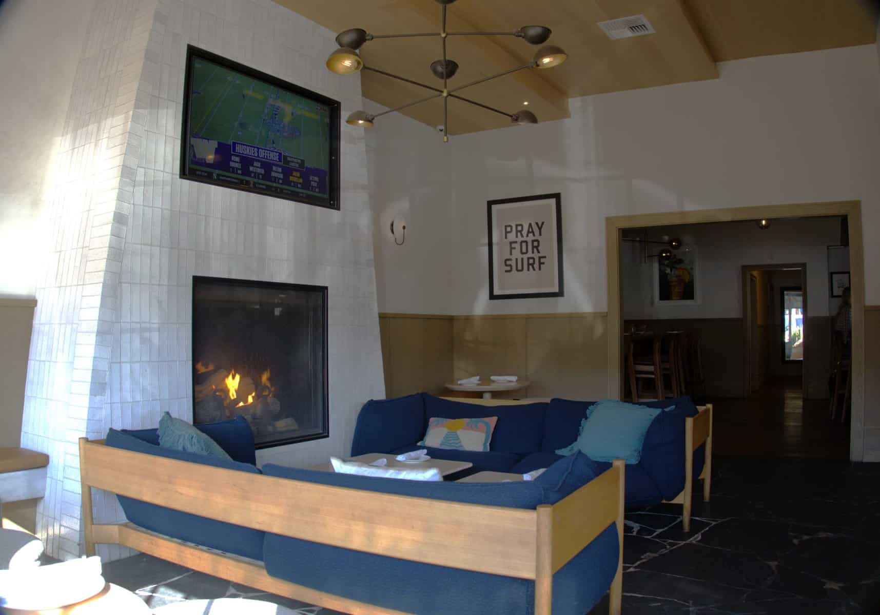 Here's a cozy lounge area with a fireplace and a big TV showing a sports game. There's a big couch with blue cushions and a sign on the wall that says "PRAY FOR SURF," which gives off a cool beach vibe. The place looks chill and comfortable for hanging out.