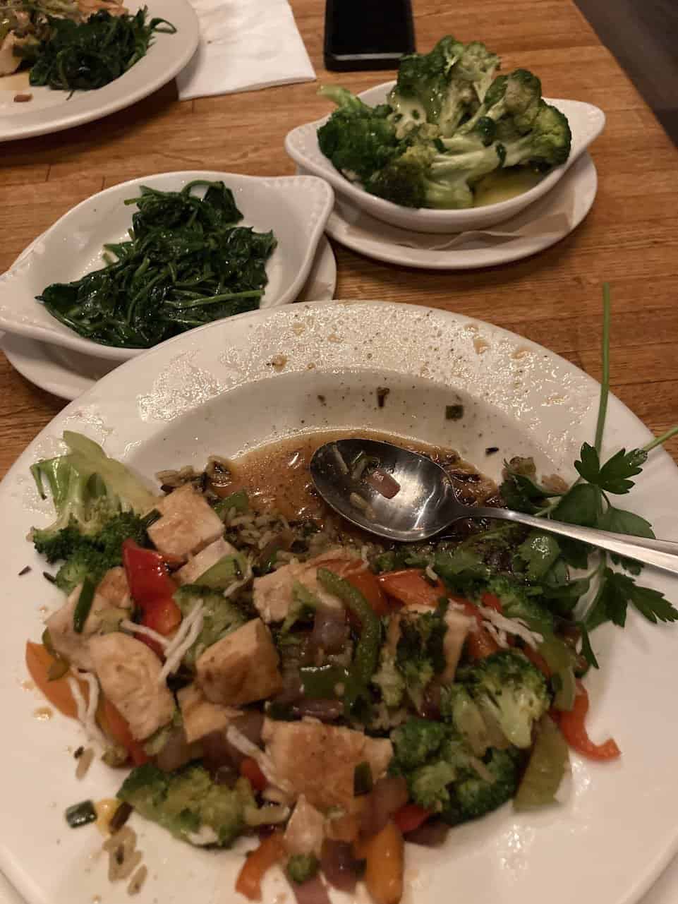 This is a photo of a meal on a wooden table. There's a white plate with a stir-fry mix of chicken and vegetables, like broccoli, carrots, and peppers, with some sauce. Behind the main dish, there are two smaller bowls, one with green spinach and the other with more broccoli. It looks like a healthy dinner option. There’s also a phone and some napkins on the table.