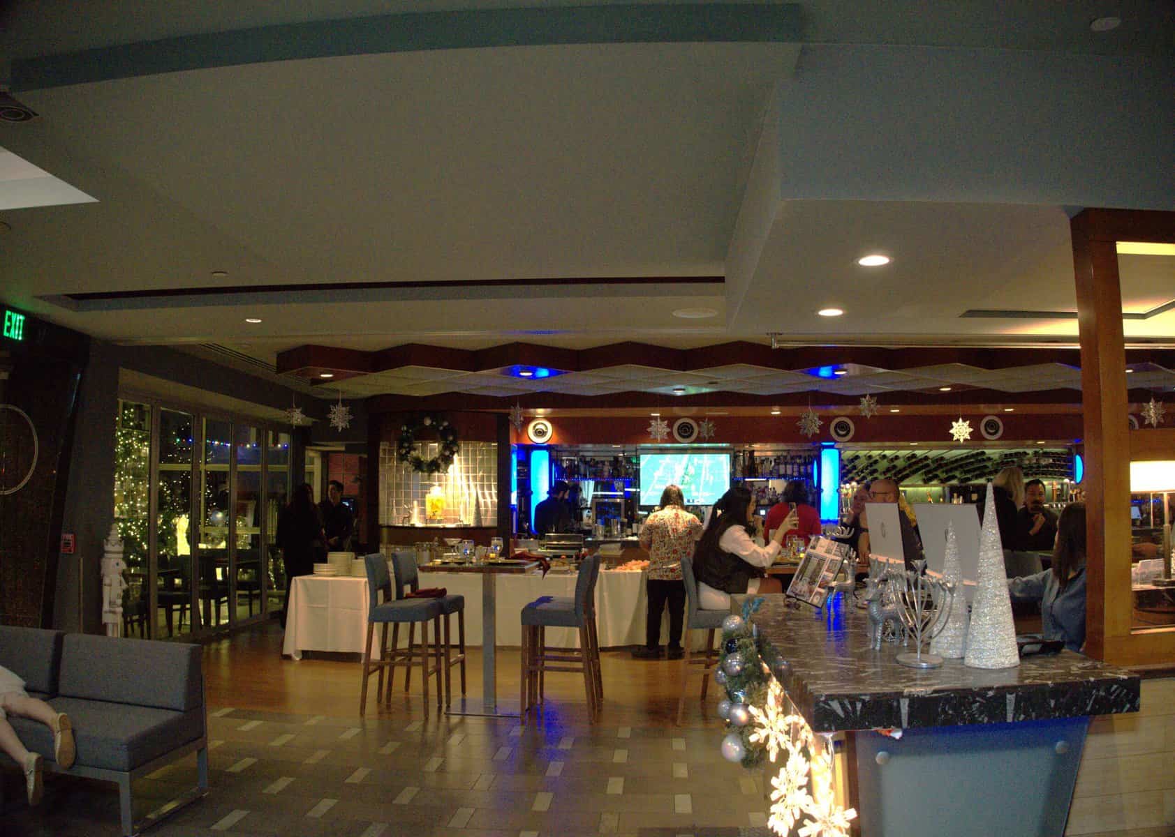 This image is of an indoor bar area that's decorated for the holidays. There are festive lights and snowflakes hanging from the ceiling, and the bar has Christmas decorations. People are sitting around, and the staff are getting ready for a holiday party or event.