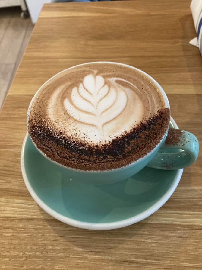 GPT
This photo shows a close-up of a hot beverage with latte art on the surface, served in a teal-colored cup that is resting on a saucer. The art looks like a leaf or a flower, made with the foam of the milk. The sprinkle of chocolate powder on top adds a nice touch to the drink. The cup is placed on a wooden table, and it seems like a cozy setting for enjoying a warm drink.