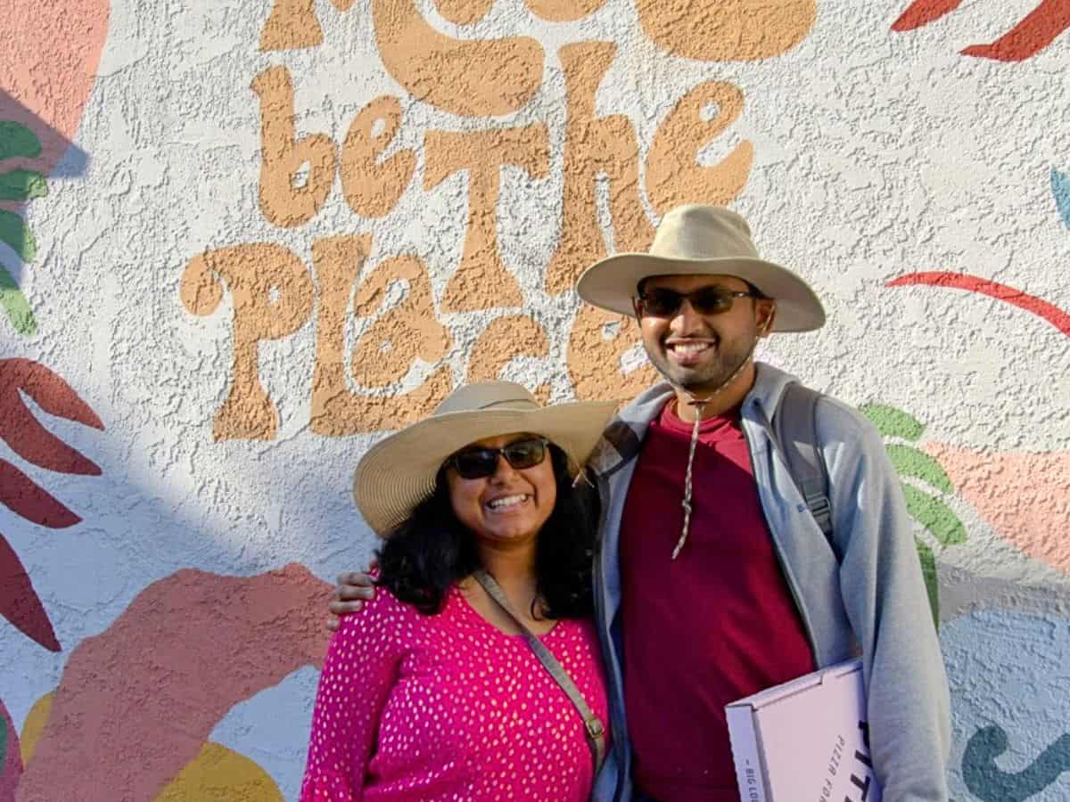 Shreeyeh and Kiran wearing sun hats and sunglasses standing in front of a colorful mural with the text "This Must Be The Place." The mural features vibrant, abstract floral designs in pink, orange, green, and blue tones.