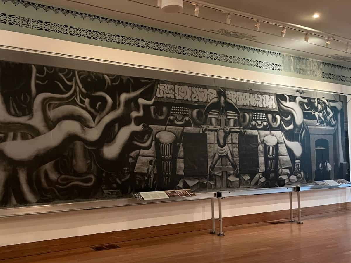 A black-and-white mural depicting bold, abstract imagery with themes of cultural history and indigenous struggles, displayed in a modern gallery space.