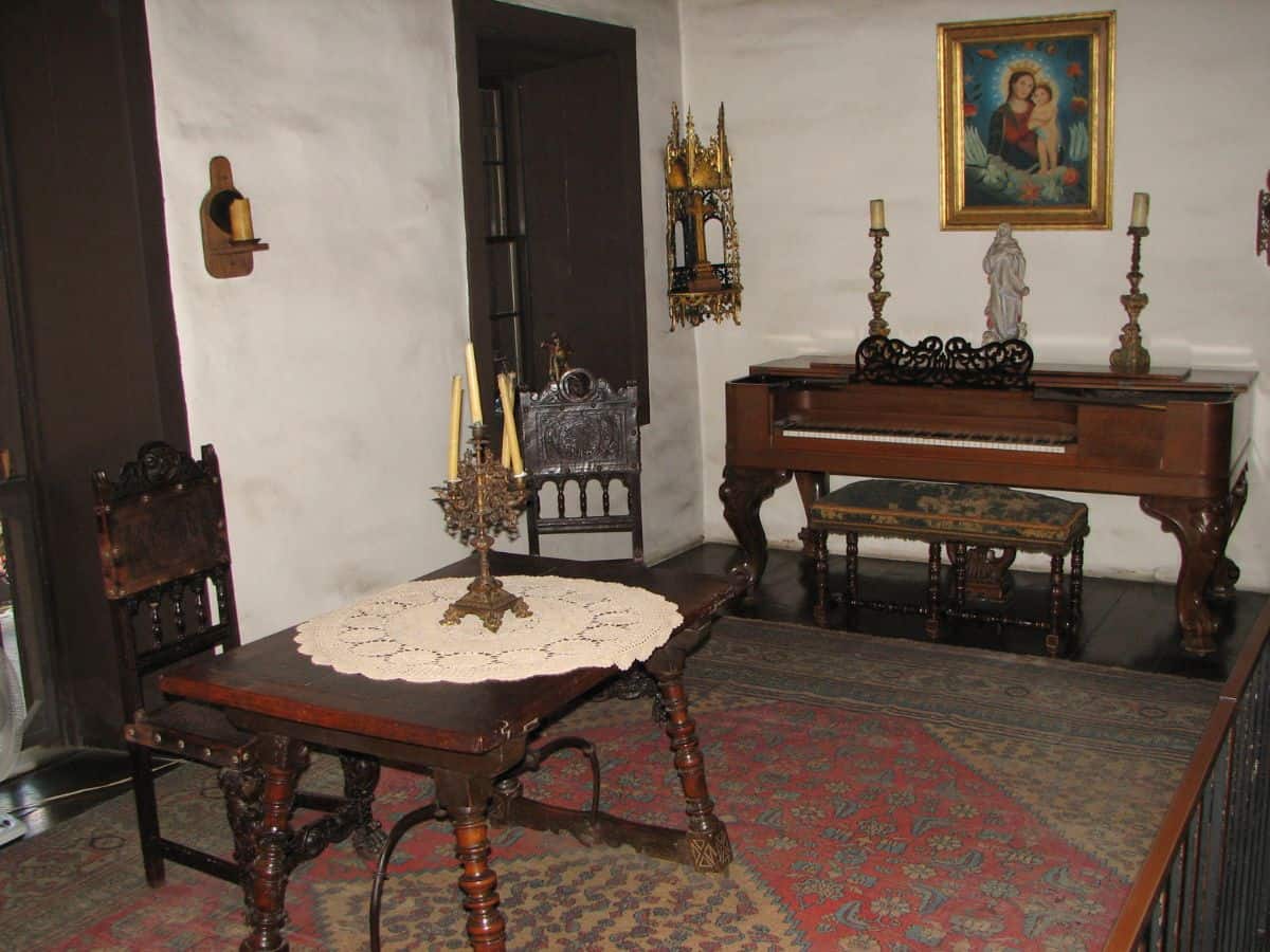 Interior of the Ávila Adobe, the oldest house in Los Angeles, featuring antique wooden furniture, a piano, and traditional Catholic artwork on the walls.