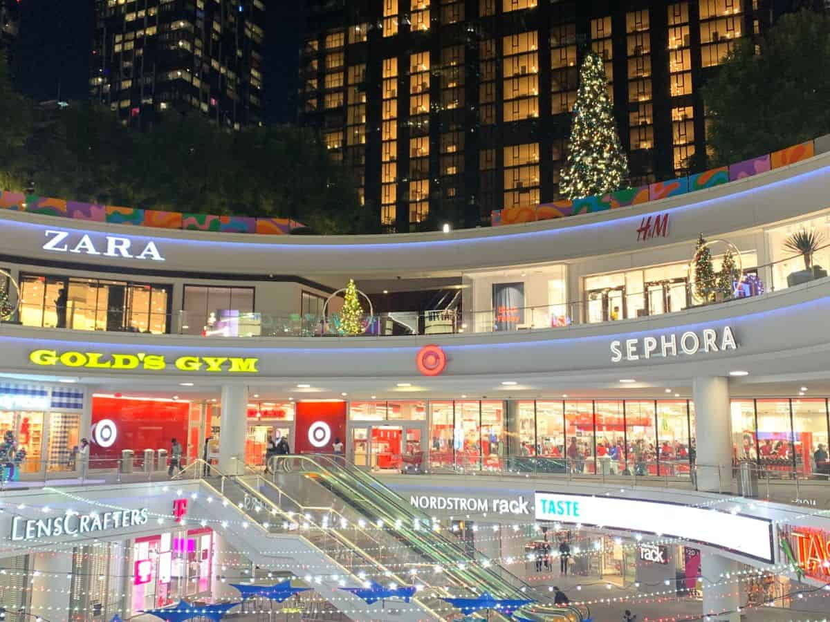 A vibrant outdoor shopping plaza at night, decorated with lights and Christmas trees, with visible stores like Zara and Sephora.