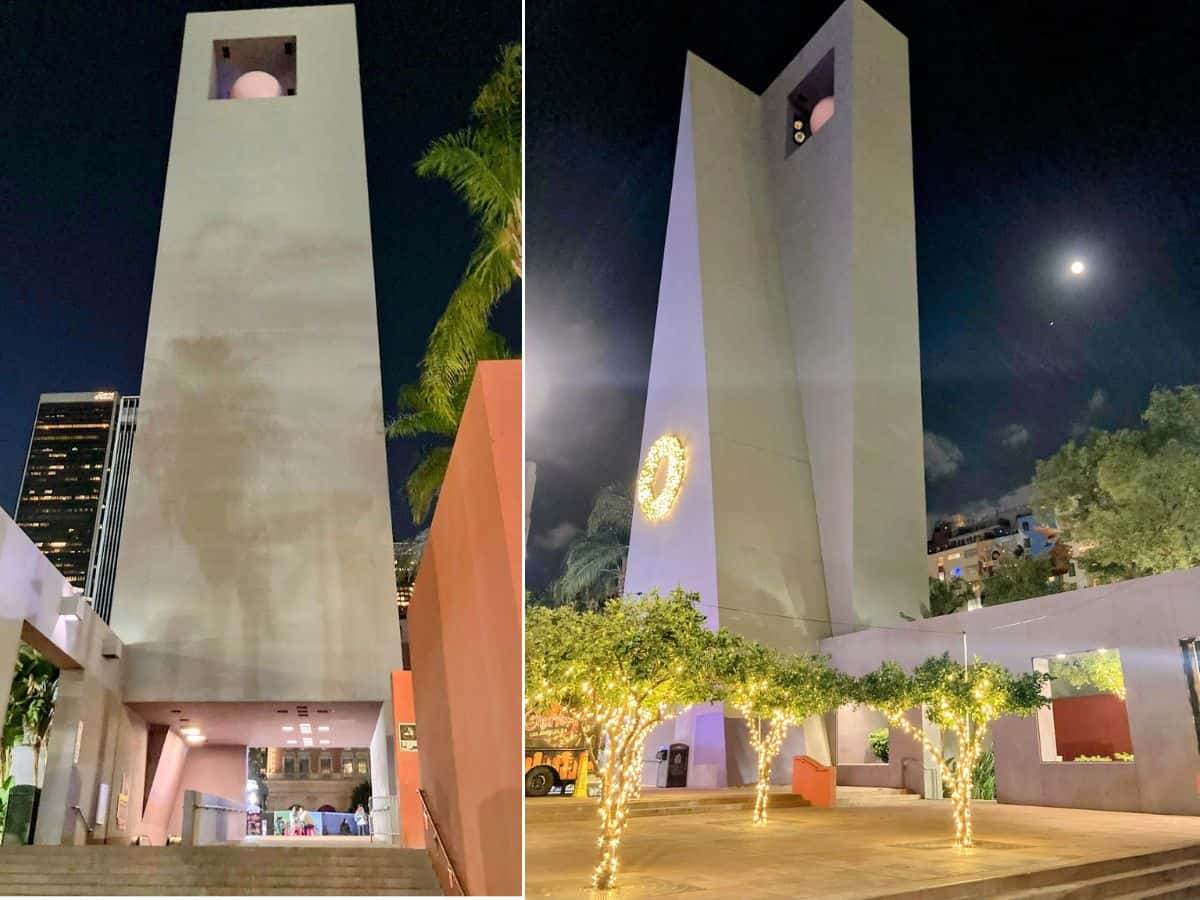 A nighttime view of the modern bell tower at Pershing Square, with illuminated string lights on nearby trees and a full moon visible in the sky.