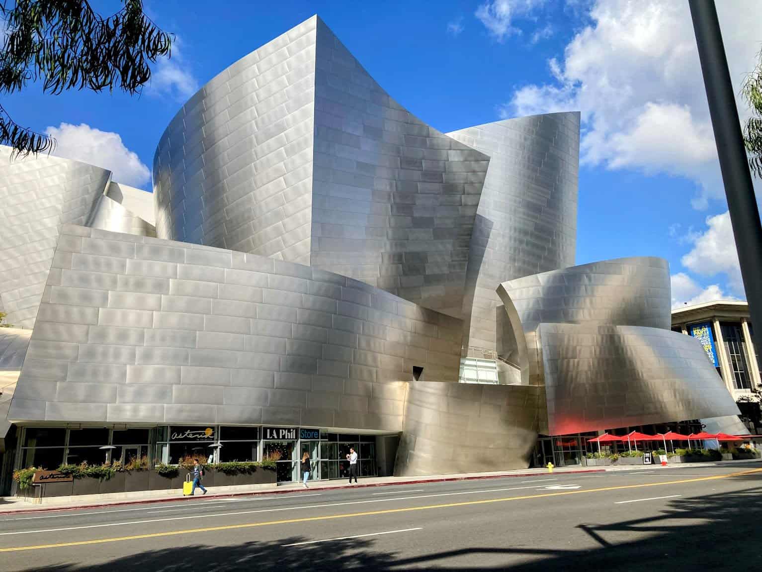 A striking view of the curved, reflective steel facade of the Walt Disney Concert Hall against a bright blue sky, surrounded by modern city buildings.