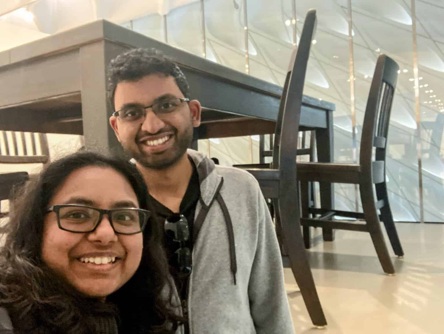 Shreeyeh and Kiran take a selfie inside an art installation featuring an oversized table and chairs, giving the impression of a whimsical, larger-than-life setting.