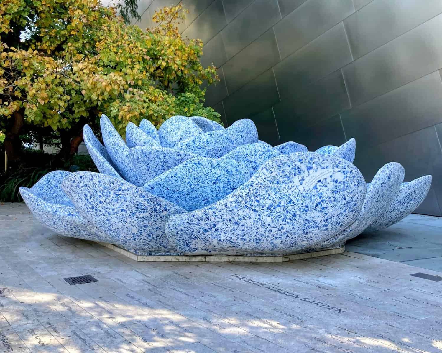 A large blue and white lotus sculpture made of mosaic tiles, situated outdoors at the Walt Disney Concert Hall, surrounded by trees and reflective architecture.
