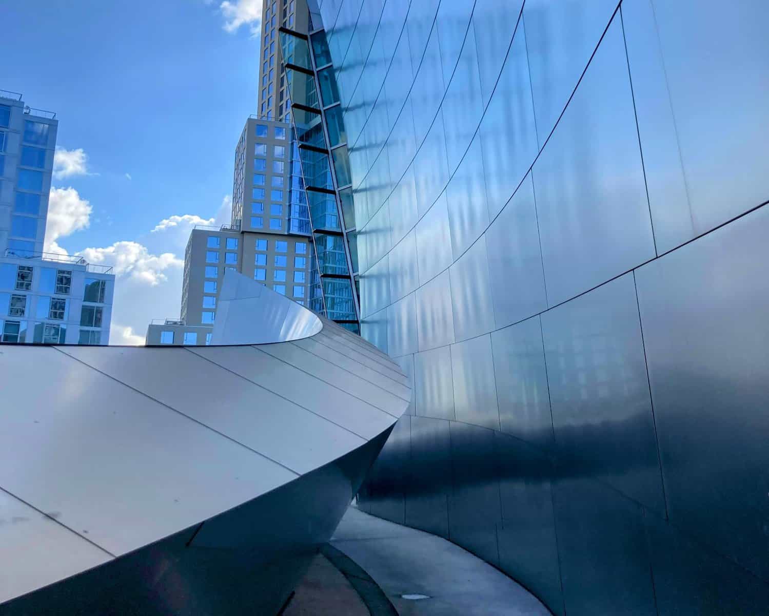 A sleek and curving silver walkway inside the Walt Disney Concert Hall, with tall, reflective surfaces and a view of nearby buildings.