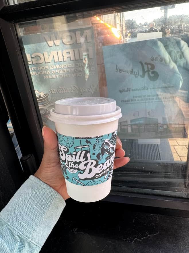 In this photo, a hand is holding a paper coffee cup with the playful words "Spill the Beans Coffee" in a teal and white pattern, a delightful find among the best things to do in Seaport Village San Diego. The backdrop reveals a reflection of the Seaport Village, hinting at the bustling ambiance outside the coffee shop
