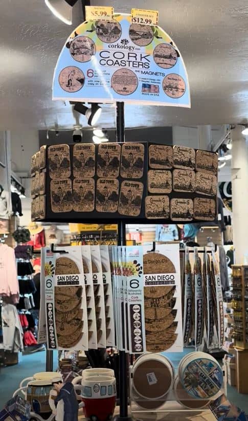 This image showcases a collection of cork coasters and magnets on display, perfect souvenirs from Seaport Village San Diego. These items are emblazoned with iconic San Diego imagery, making them great keepsakes or gifts. Shoppers looking for a piece of San Diego to take home will find these items, along with other local memorabilia, in the shops of Seaport Village, which is among the best things to do in the area. The display is set against a backdrop of other tourist-friendly goods, highlighting the vibrant shopping experience at Seaport Village