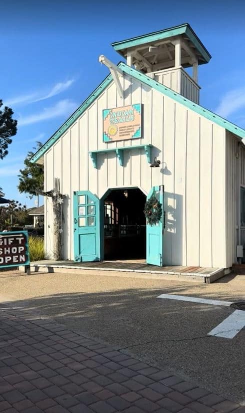 In this sunny scene, the Indian Trails gift shop stands out with its distinctive pale blue façade and welcoming teal doors that are propped open, inviting in any curious visitor. Above the entrance, a charming sign adorned with Native American-inspired designs beckons shoppers to discover the treasures inside. This shop is a wonderful stop for those looking for unique souvenirs and is part of the vibrant shopping experience at Seaport Village, making it one of the best things to do in San Diego. Its traditional structure, complete with a rustic wooden exterior and a small tower feature, adds to the quaint charm of the village, set against the clear blue sky—a perfect memento of San Diego's rich heritage and culture