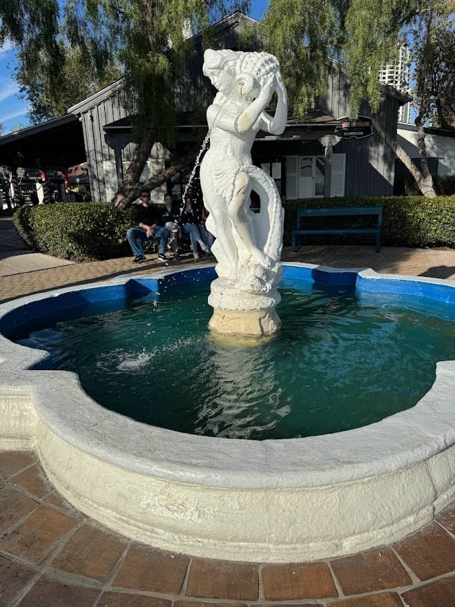This image features a whimsical white seahorse fountain rising from the center of a bright blue tiled circular pool, one of the best things to do in Seaport Village San Diego. The water cascades gently around the statue, adding a serene soundtrack to the bustling village atmosphere. In the background, visitors are seated on benches beneath the shade of lush trees, adjacent to charming shops with rustic wooden facades. It's a peaceful spot for relaxation and an Instagram-worthy photo opportunity in the heart of Seaport Village.