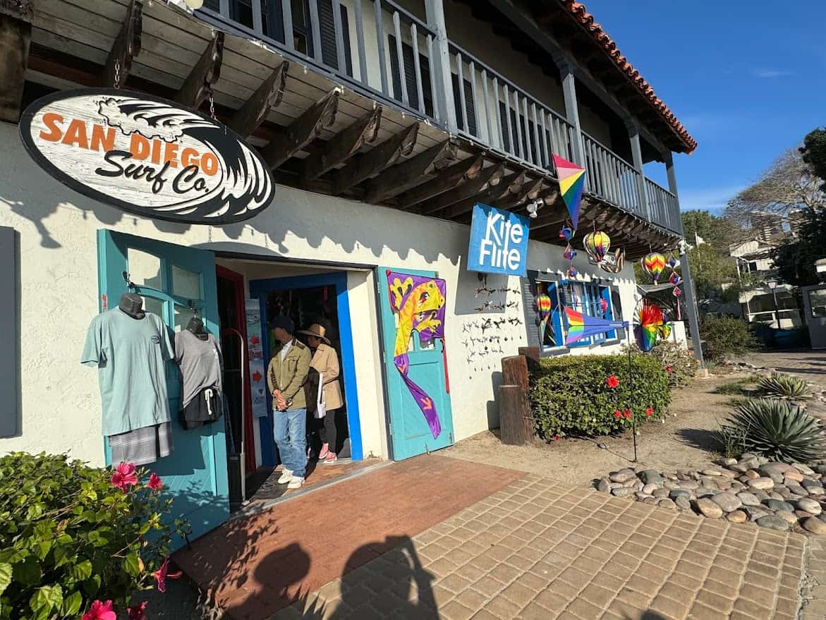 In this photo, we're looking at the sunlit entrance of San Diego Surf Co., a shop that captures the essence of the city's surf culture. With its iconic sign and selection of casual surf apparel displayed outside, it invites both locals and visitors to explore its collection. Next door, the vibrant colors of kites and wind ornaments at the Kite Flite shop add a playful touch, highlighting the fun and diverse shopping experience that makes visiting Seaport Village one of the best things to do in San Diego. The shop fronts with their unique offerings sit under a traditional Spanish balcony, surrounded by bright flowers and a cobblestone path, enhancing the charming coastal vibe of the village.