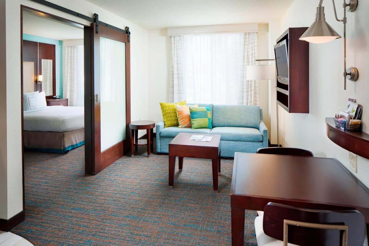 Inviting hotel suite featuring a partitioned bedroom, a turquoise sofa with vibrant pillows, and a neat work desk with contemporary amenities.