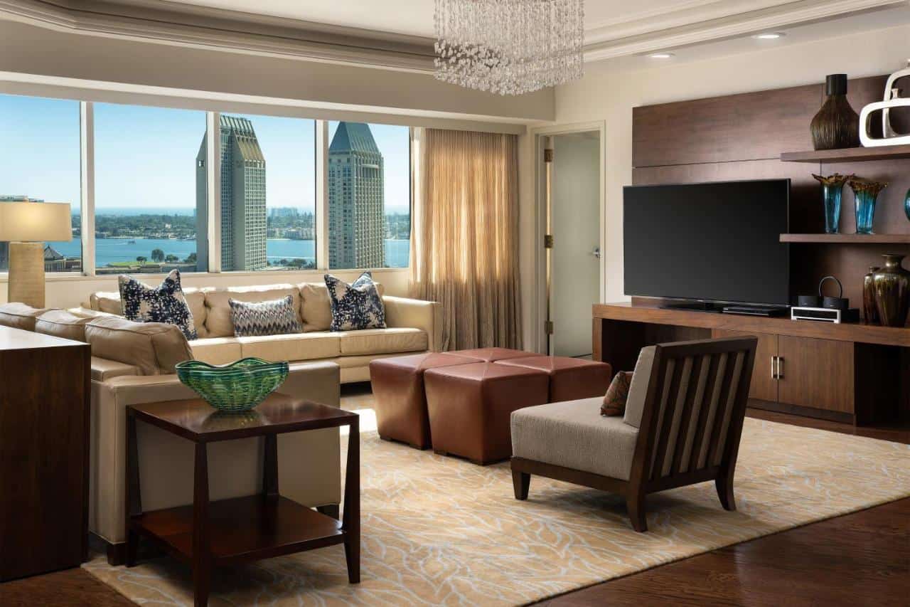Luxurious hotel living area with a large beige sectional sofa adorned with decorative pillows, wood and leather accents, and a grand window offering a sweeping view of the San Diego bay and skyline.