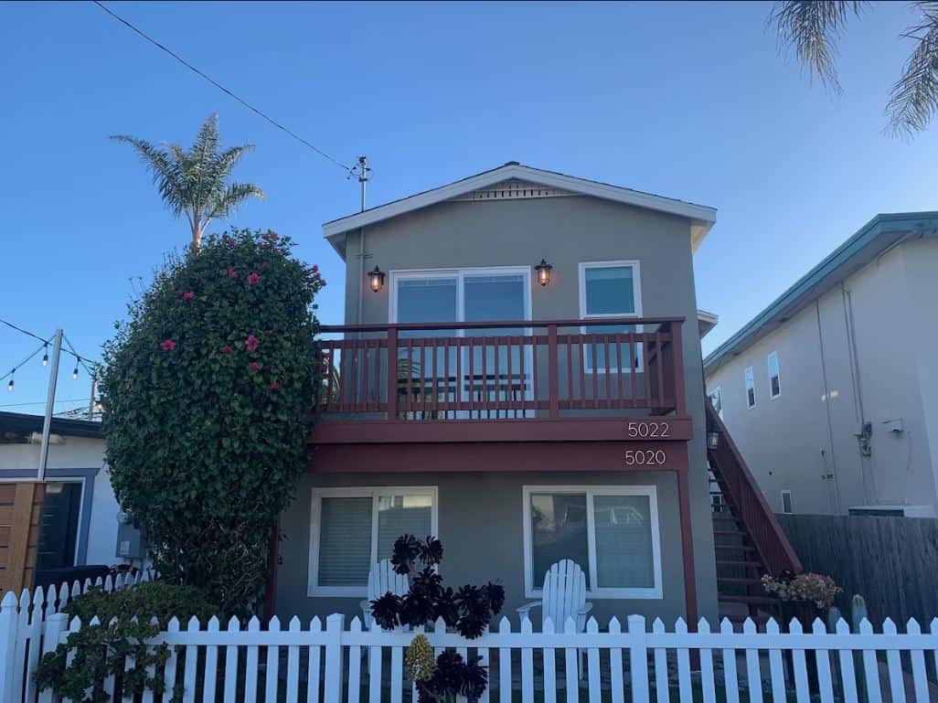 Exterior view of a charming, two-story Airbnb rental in Ocean Beach San Diego with a spacious balcony adorned with string lights and surrounded by lush greenery