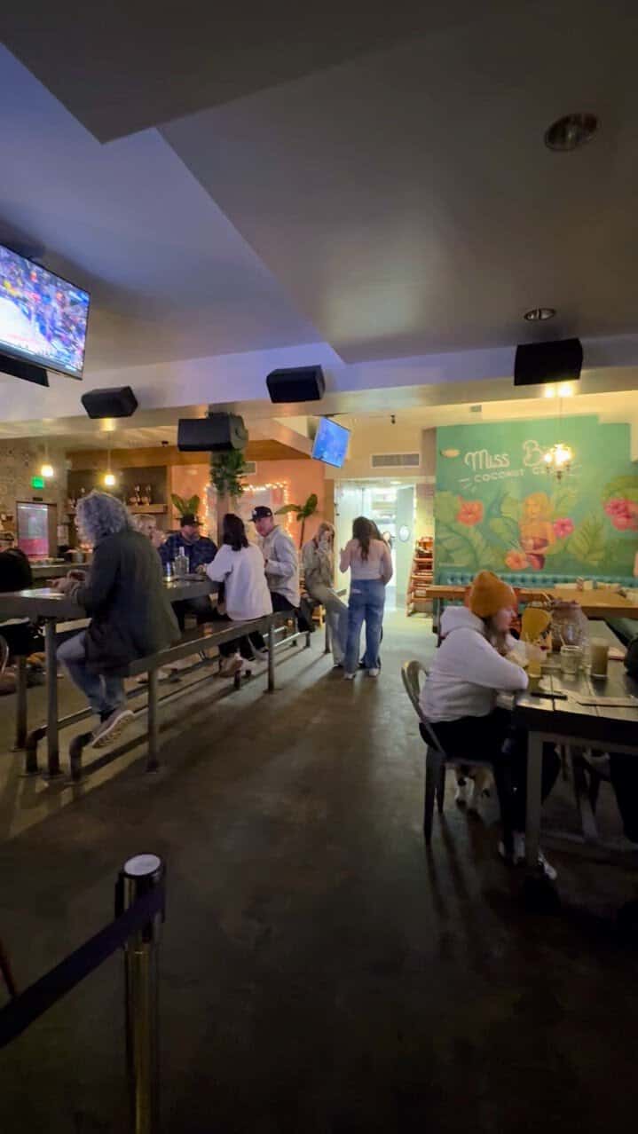 Interior of Miss B's Coconut Club, one of the best bars in Mission Beach, San Diego, showing a lively atmosphere with patrons seated at long tables and watching sports on multiple TVs. The decor includes tropical-themed murals and warm lighting.