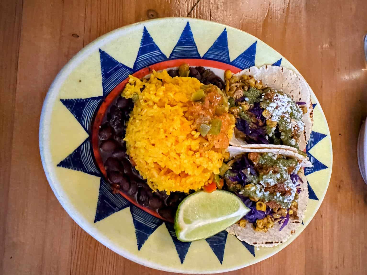 A colorful plate featuring vibrant yellow rice, black beans, and two tacos filled with seasoned vegetables, purple cabbage, and sprinkled cheese, served with a lime wedge. The plate has a bold geometric pattern with blue and yellow accents, enhancing the vibrant presentation of the dish.