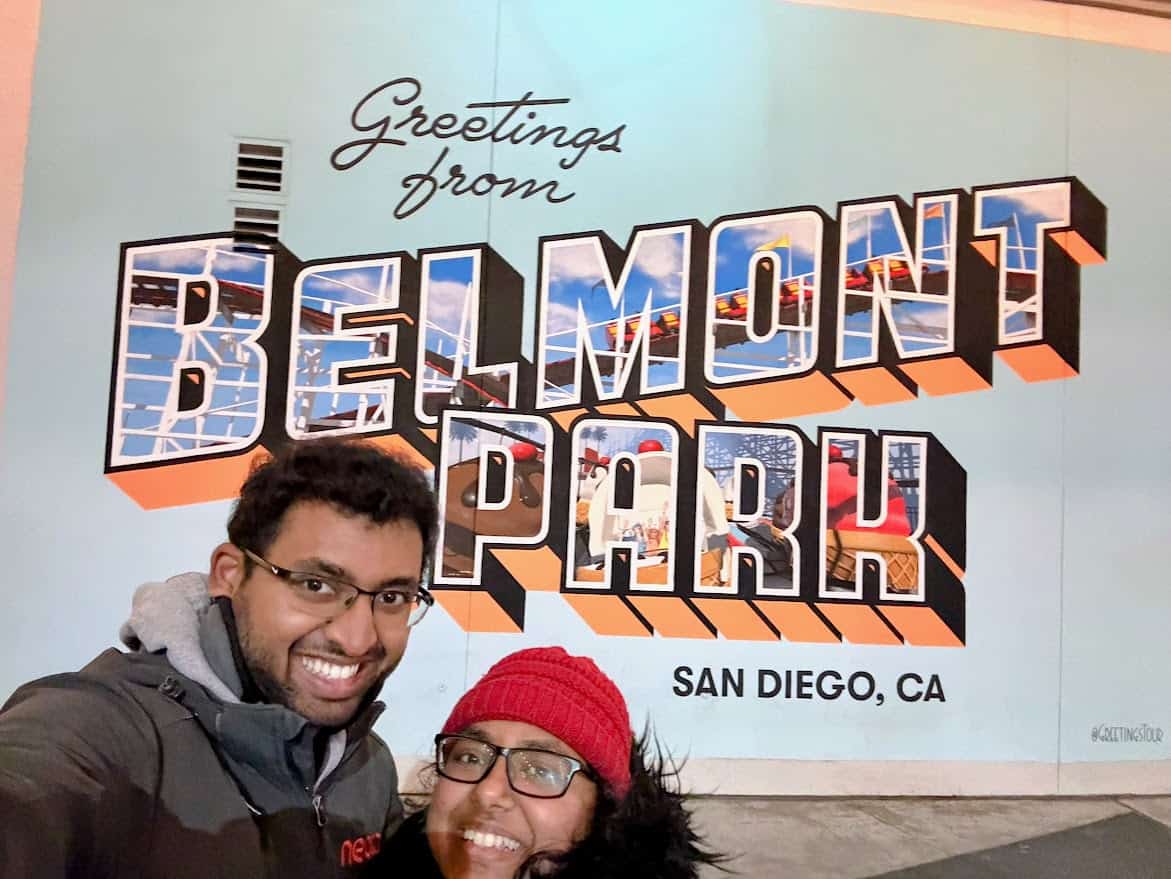 A cheerful mural reading "Greetings from Belmont Park" in San Diego, CA, with a couple smiling in front of it, capturing a perfect photo opportunity and one of the fun things to do in Belmont Park.