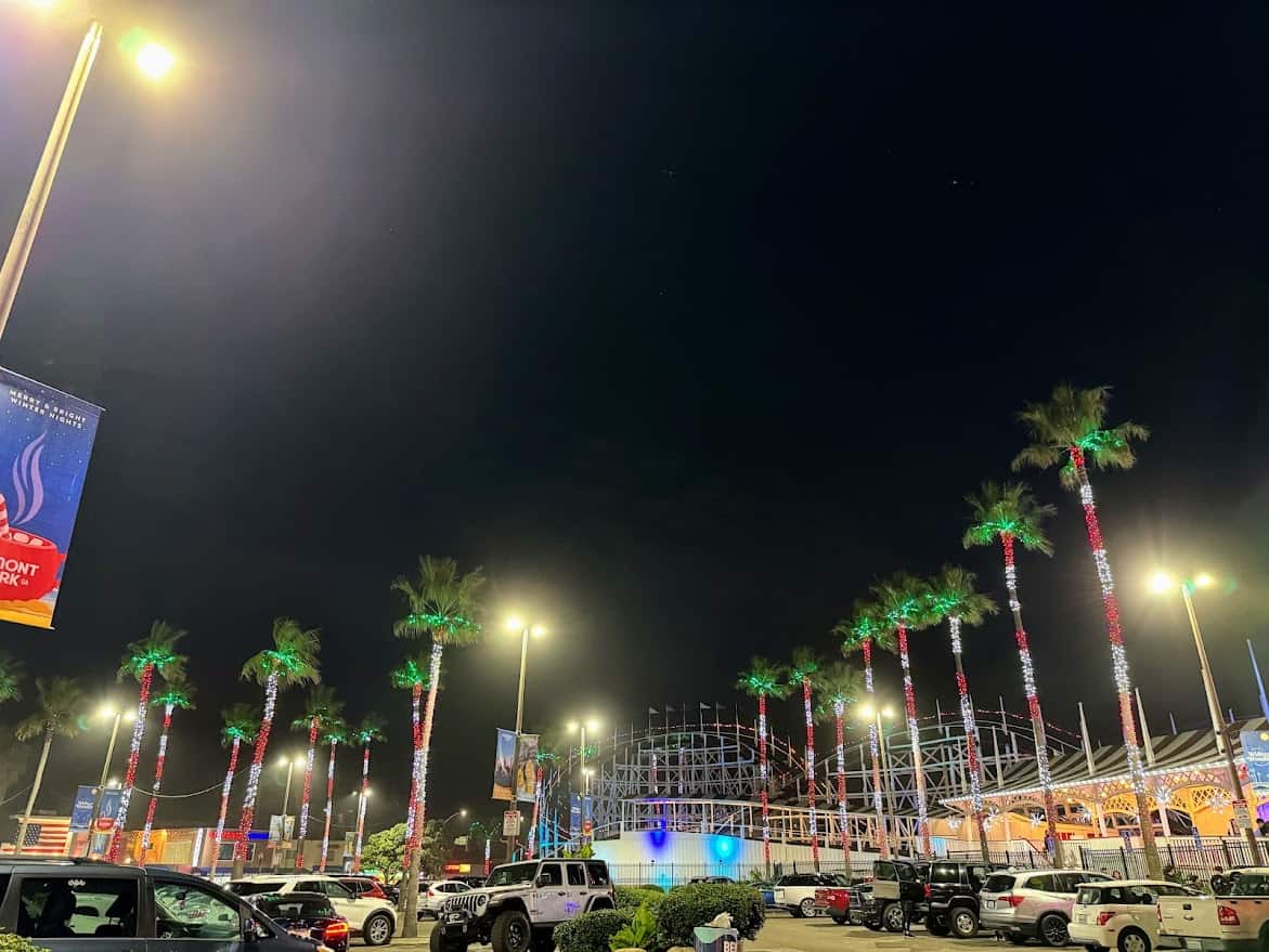 A nighttime view of the Belmont Park parking lot, with palm trees wrapped in festive lights and the iconic Giant Dipper roller coaster in the background, showcasing one of the many things to do in Belmont Park, Mission Beach.