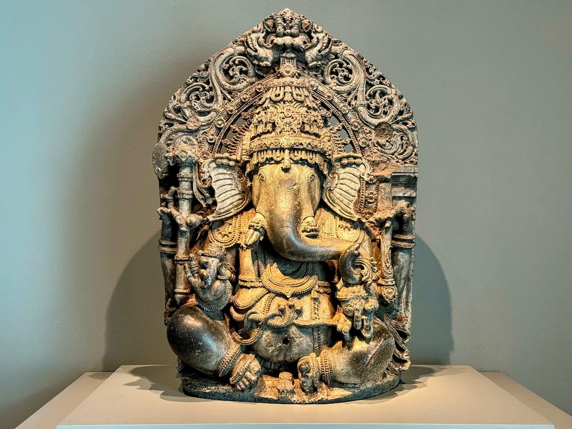 A detailed sculpture of the Hindu deity Ganesha, displayed in at he Asian Art museum. The sculpture is intricately carved with ornate decorations surrounding the figure, showing the deity with an elephant head and multiple arms. The lighting highlights the stone texture, giving it a majestic appearance against the plain background.