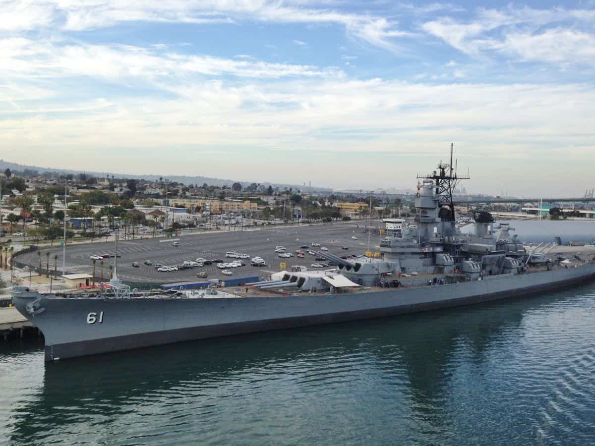 A large naval ship, USS Iowa docked at the port in Los Angeles, with the number "61" on its bow. The battleship is a popular historic attraction, with a view of the surrounding cityscape in the background. Cars are visible in a nearby parking lot, and the sky above is partially cloudy, creating a peaceful scene by the waterfront.
