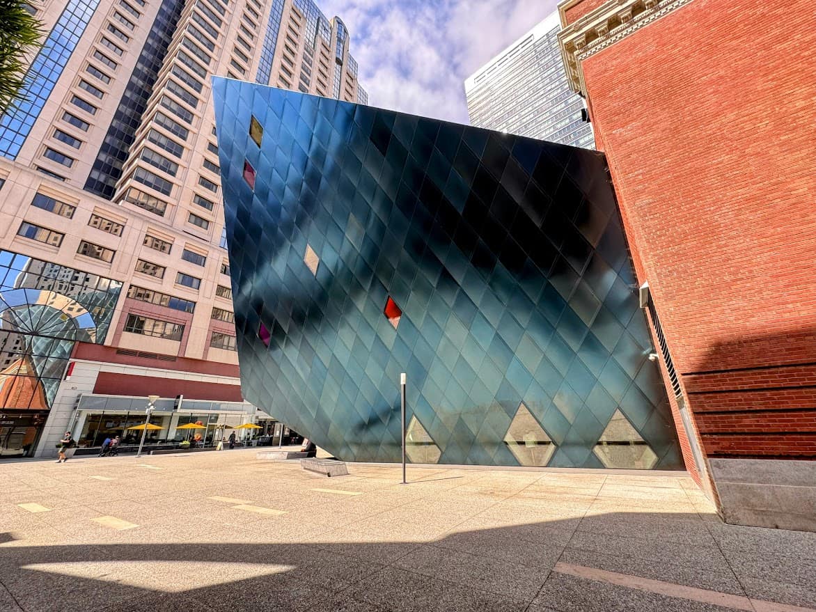 The image shows the Contemporary Jewish Museum (CJM) in San Francisco, with its distinctive modern architecture. The building features a striking geometric design, clad in blueish metallic panels, creating a dramatic contrast with the surrounding brick structure and nearby high-rise buildings. Colorful windows are scattered across the angled walls, reflecting light and adding a dynamic element to the scene.