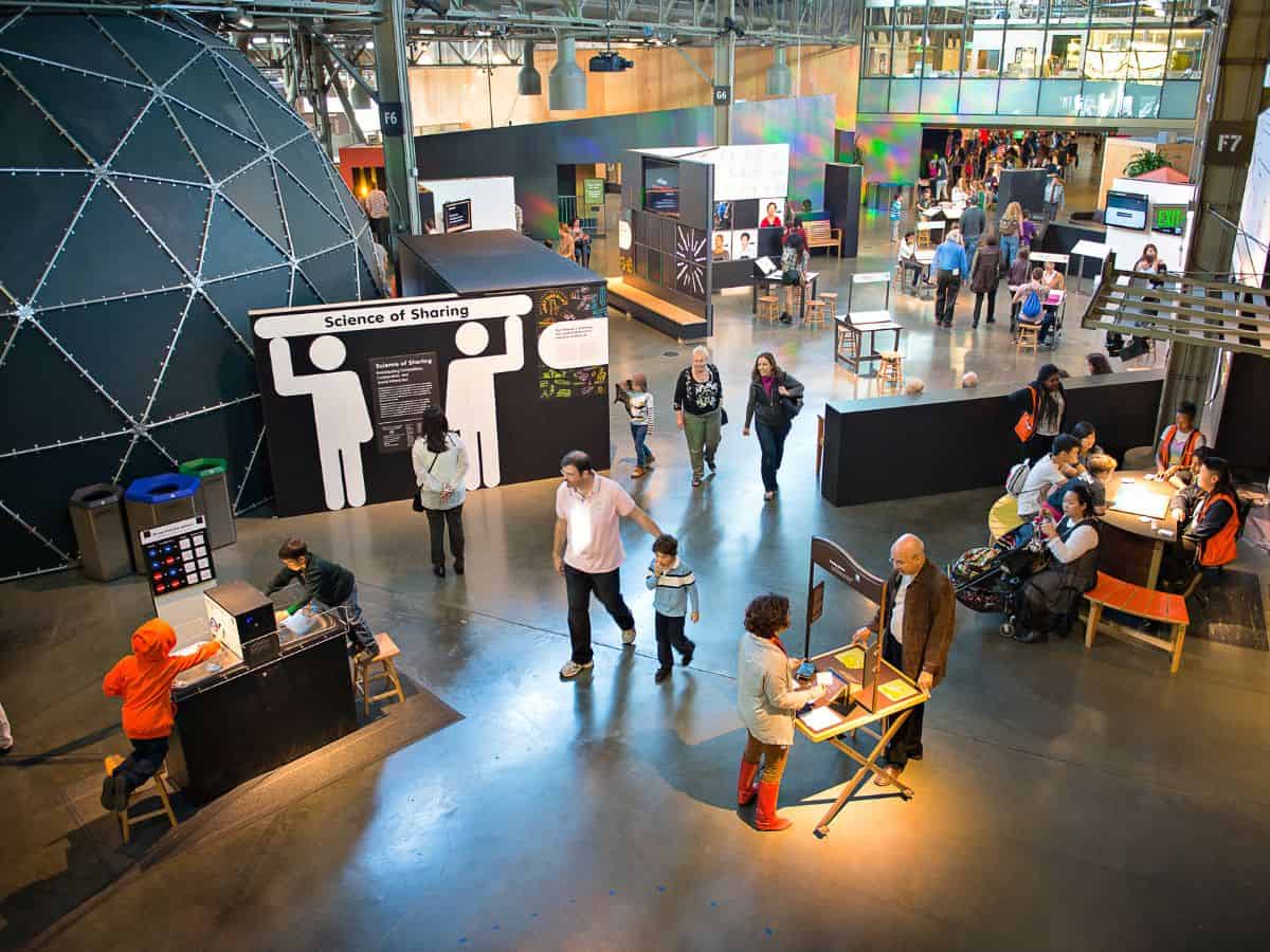 A lively interior scene at the Exploratorium in San Francisco, showcasing interactive exhibits and visitors engaging in hands-on science activities. People are exploring the "Science of Sharing" exhibit and other stations. Children and adults are actively participating in the displays. There's a large, geometric dome structure in the background.