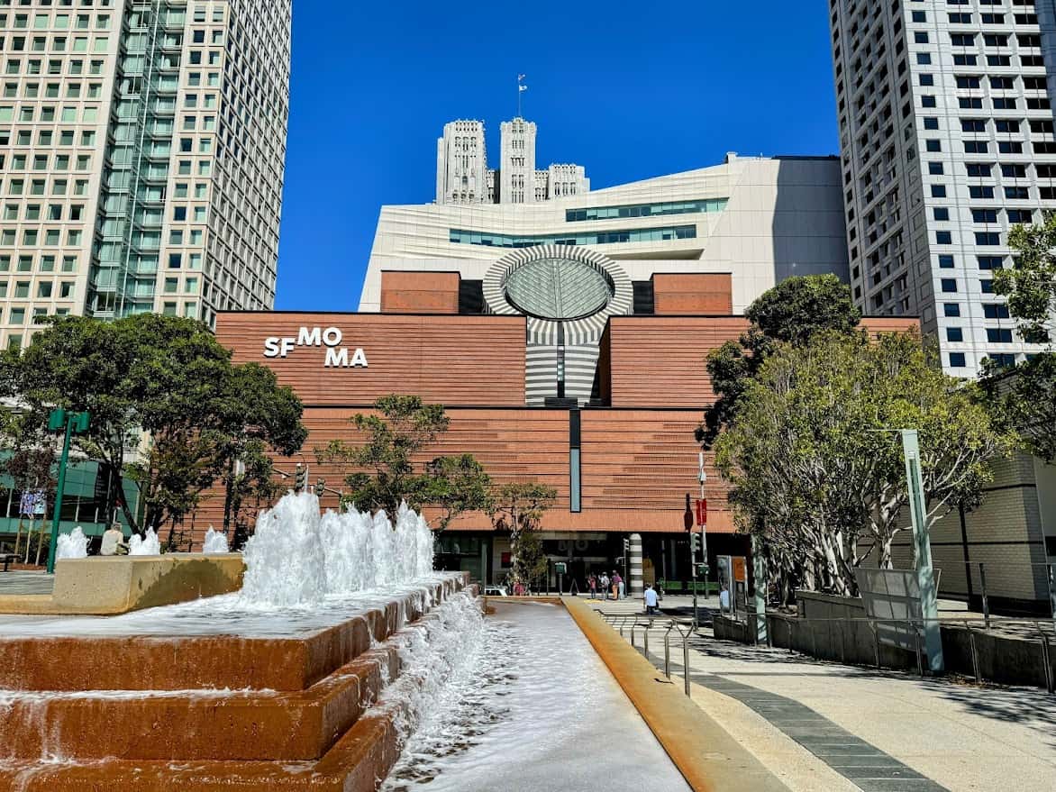 A view of the San Francisco Museum of Modern Art (SFMOMA) building, with a sleek, modern architectural design. The image also includes a vibrant water fountain in the foreground, adding dynamic movement to the scene. Tall city buildings surround the museum, showcasing the urban setting.
