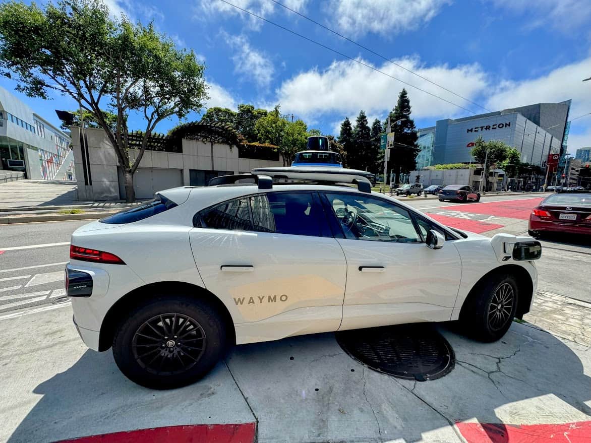 A Waymo self-driving car is parked on the side of a sunny street in an urban area. The white electric vehicle is equipped with various sensors on the roof and sides, used for autonomous driving.